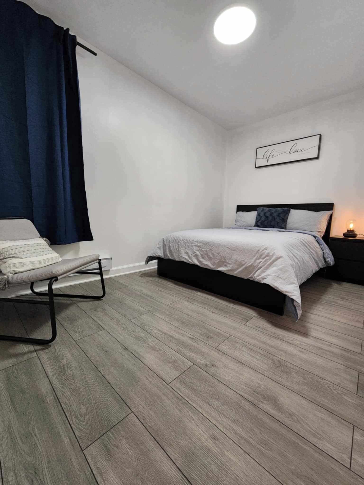 Une chambre à coucher avec un lit, une table de chevet avec une lampe, une chaise et un mur orné d’une affiche.