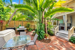 A patio with a glass table and chairs, surrounded by lush greenery and a wooden fence. - Turtle Nest Condo + Pool / 2nd Floor (Key West)
