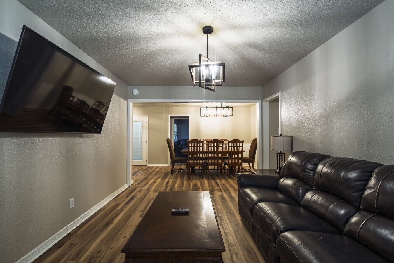 A living room with a leather sofa, a dining table with chairs, a TV mounted on the wall, and a chandelier.