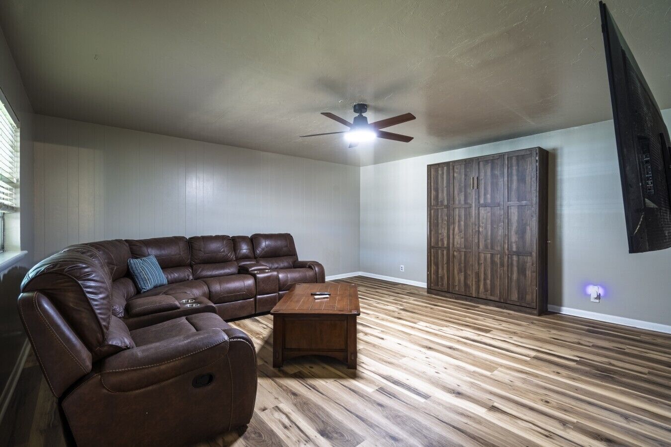 A room with a brown leather sectional sofa, a wooden coffee table, a ceiling fan, and a wooden wardrobe.