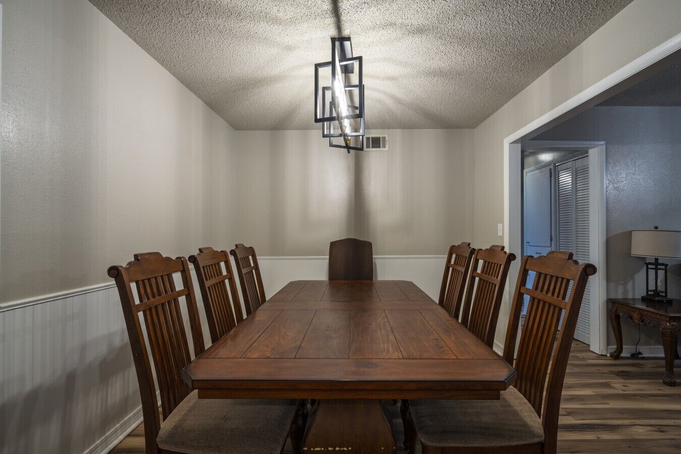 A dining room with a rectangular wooden table surrounded by six chairs, a ceiling light fixture, and a door leading to another room.