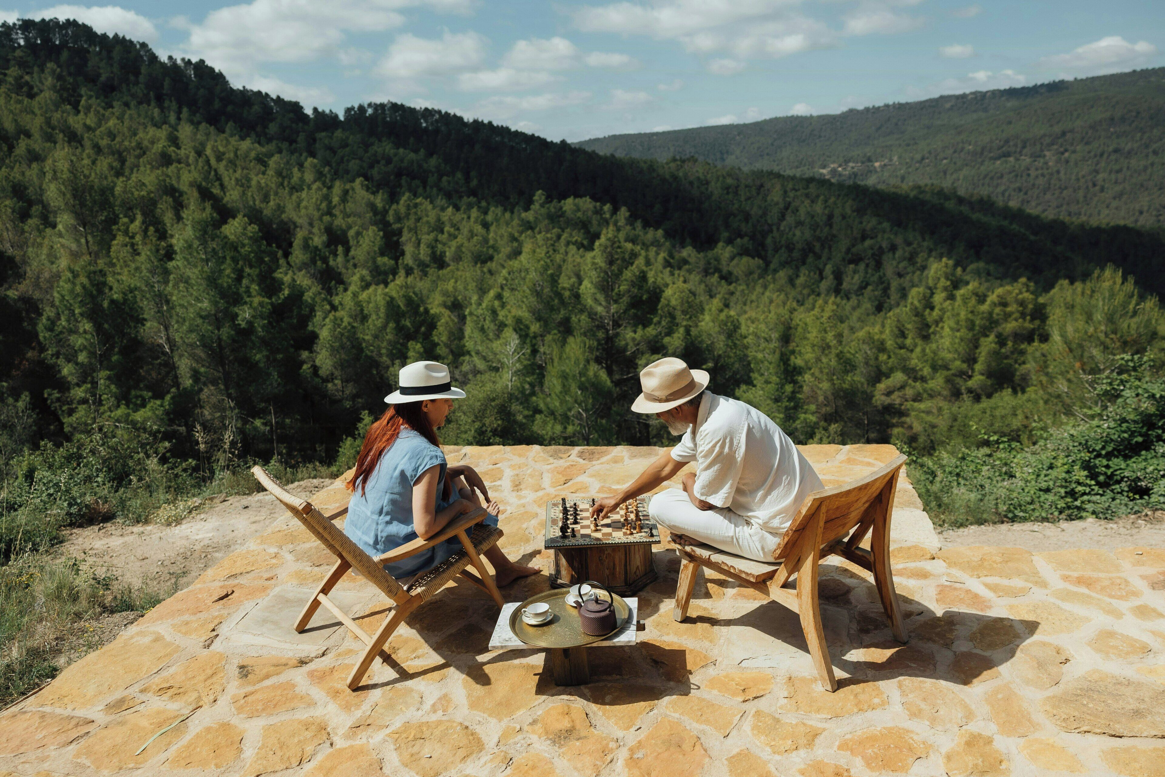 Dos personas jugando ajedrez sobre una mesa de piedra al aire libre, con vista a un bosque.