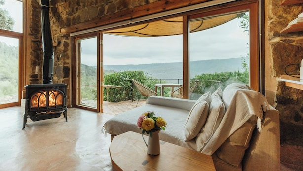 A cozy room with a stone wall, a large window with a view, a wooden sofa, and a fireplace.