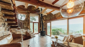 A rustic interior with stone walls, wooden beams, a fireplace, and large windows offering a view of the outdoors.