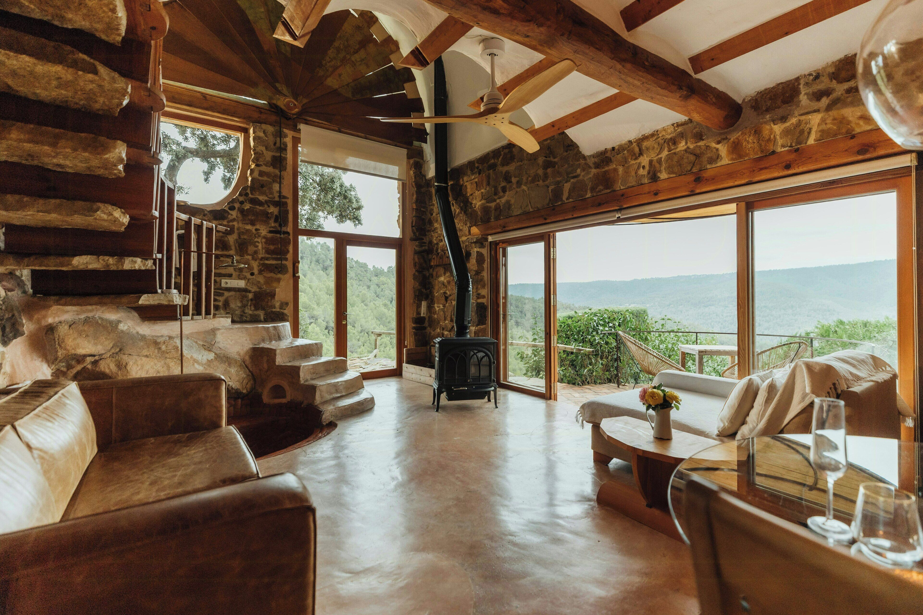Un intérieur rustique avec des murs en pierre, des poutres en bois, une cheminée et de grandes fenêtres donnant sur l’extérieur.