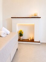 Un dormitorio con chimenea de ladrillo blanco, una toalla blanca enrollada sobre la cama, un jarrón con girasoles y dos velas en la repisa.