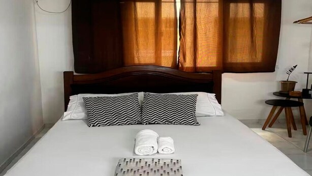 Una cama bien hecha con cabecera de madera, dos almohadas estampadas y toallas enrolladas sobre la cama.
