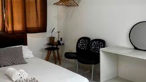 Habitación de hotel con una cama, un escritorio, dos sillas, una mesita con una planta y un perchero.