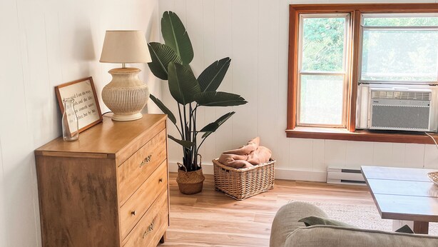 Une pièce comprenant une commode en bois, une lampe, une plante en pot, un panier en osier et une fenêtre avec un climatiseur.