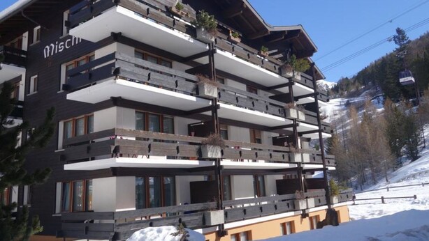 Un hotel a più piani con balconi, una seggiovia sullo sfondo e neve a terra.