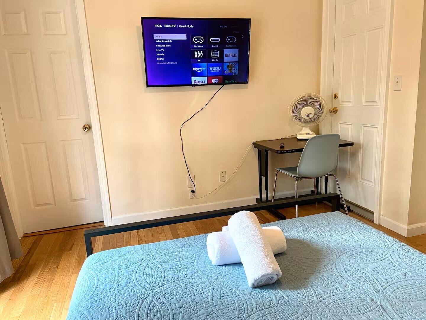 Ein Zimmer mit einem Bett, einem Schreibtisch, einem Stuhl, einem an der Wand montierten Fernseher und einem Ventilator.