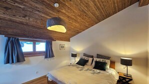 Makuuhuoneessa on puinen katto, valkoisilla lakanoiden peitetty sänky, kaksi yöpöytää, joissa on lamput, ja ikkuna, jossa on verhot.
