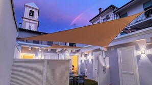 Ein überdachter Patio mit Tisch und Stühlen, ein weißer Zaun und im Hintergrund ein Gebäude mit einem Glockenturm.