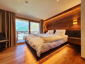 Ein modernes Schlafzimmer mit einem großen Bett, einem Holz-Kopfteil und Blick auf eine gebirgige Landschaft durch das Fenster.