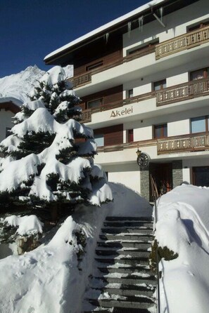 A hotel with a snow-covered entrance, a staircase leading up, and a mountain in the background. - Akelei Appartement/Fewo 4-Bettwohnung by Interhome (Saas-Fee)