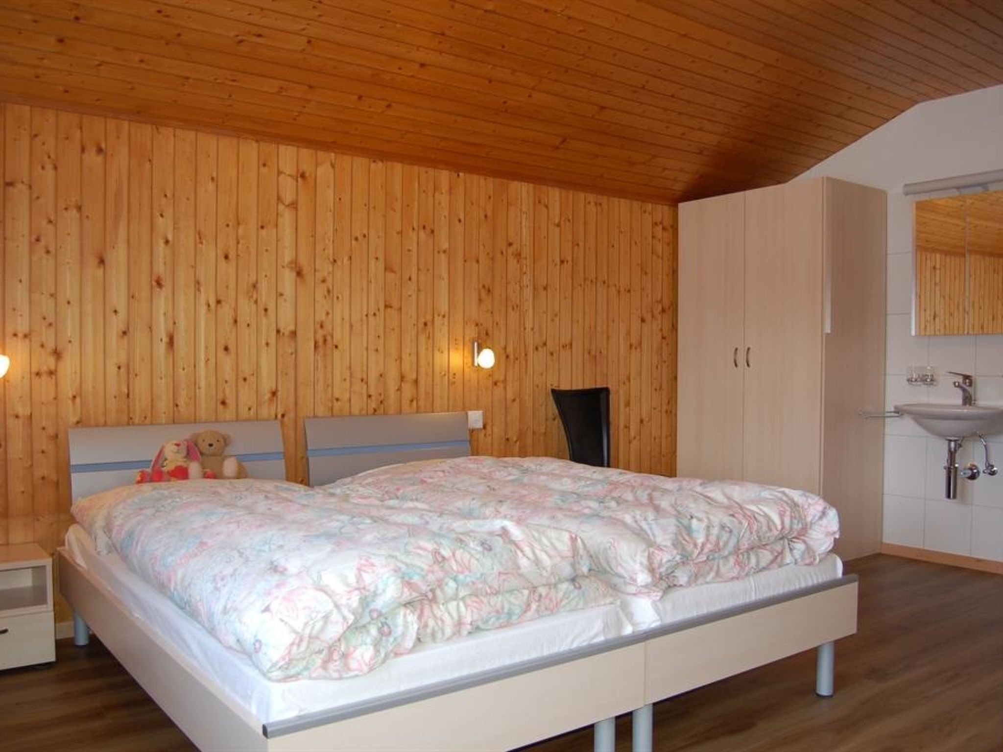 Quarto com teto de madeira, cama com colcha florida, mesa de cabeceira, cômoda e banheiro com pia e espelho.