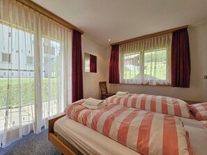 A bedroom with two beds, large windows with curtains, a view of greenery outside, and a small table with a chair. - Dominic B1 03 Appartement/Fewo 6-Bettwohnung by Interhome (Saas-Fee)