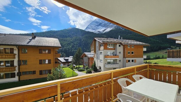 Ein Balkon mit Holzgeländern, einem Tisch und Stühlen mit Blick auf einen Berg und Gebäude.