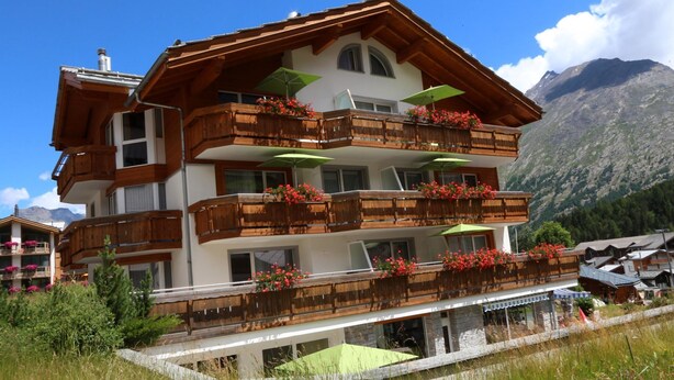 Un hotel de varios pisos con balcones de madera, mobiliario exterior y un paisaje montañoso de fondo.