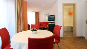 Comedor con una mesa redonda, sillas rojas, un televisor sobre un mueble bajo y un baño a la vista a través de una puerta abierta.
