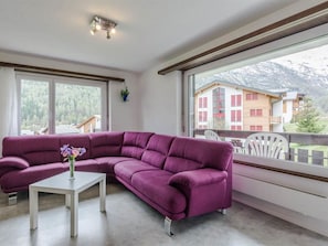 A modern living room with a purple sectional sofa, a white coffee table, and a view of a mountain landscape through large windows. - Alouette Appartement/Fewo 6-Bettwohnung Nr. 13 by Interhome (Saas-Fee)