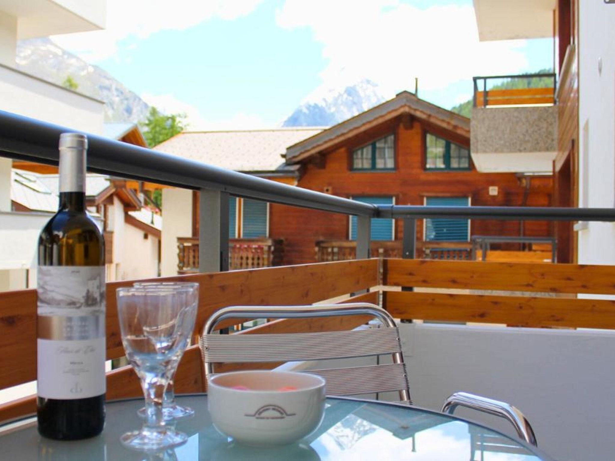 Ein Tisch mit einer Weinflasche, einem Glas und einer Schüssel, mit Blick auf eine Holzterrasse und eine Berglandschaft.