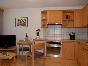 A compact kitchen with wooden cabinets, a built-in oven, a coffee maker, and a dining area with a table and chairs. - Swiss Chalet Appartement/Fewo 2-Bettwohnung Nr. 7 by Interhome (Saas-Fee)