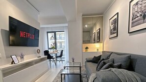 A modern living room with a grey sofa, a coffee table, a TV mounted on the wall, and a dining area with chairs and a table.