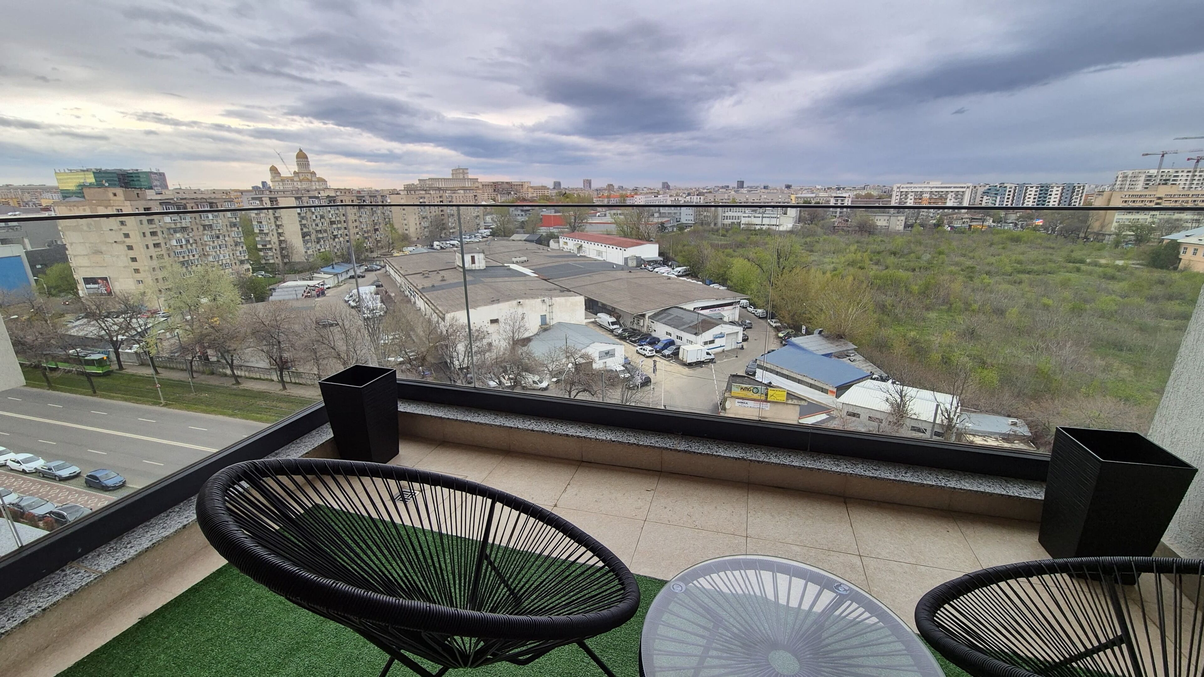 Un balcón con dos sillas negras y una mesa, con vista a un paisaje urbano con edificios y una grúa de construcción.