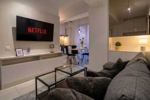 Ein modernes Wohnzimmer mit Flachbildfernseher, einer Couch, einem Glastisch und einer Küche im Hintergrund.