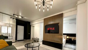A modern living room with a large flat-screen TV displaying "NETFLIX," a glass coffee table, a sofa with a yellow cushion, and a minimalist design with wooden accents.
