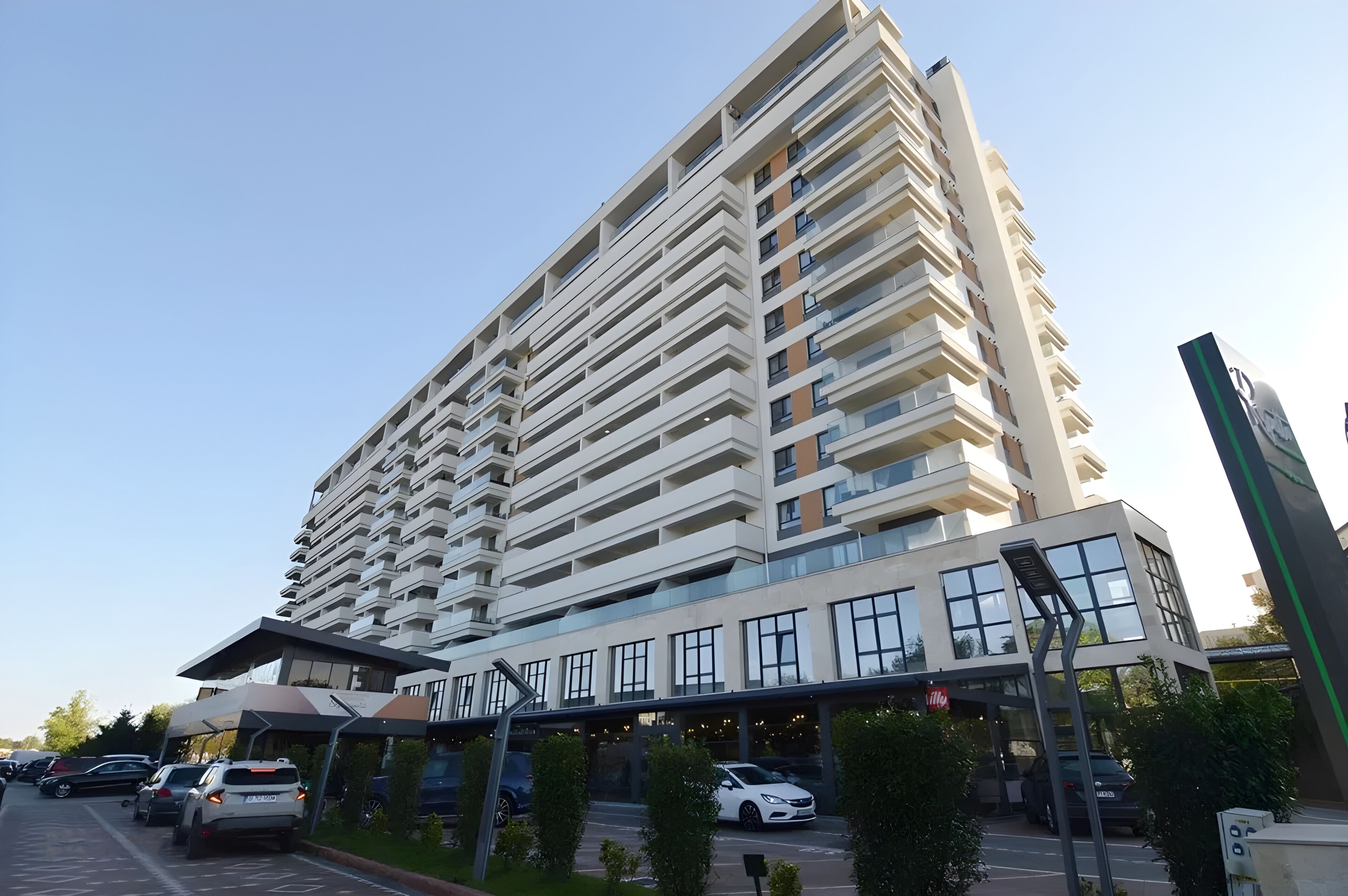 Een hoog gebouw met meerdere balkons, een overdekte ingang en een parkeerplaats met auto's.