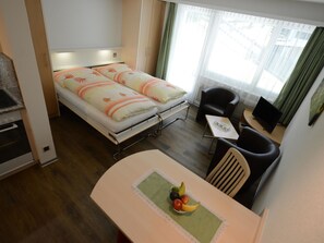Habitación de hotel compacta con una cama, una pequeña mesa de comedor con fruta, una silla y un televisor.