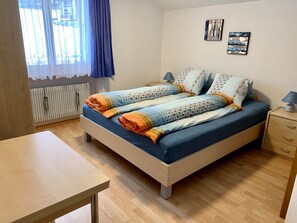 A bedroom with a bed, a bedside table, a lamp, a radiator, and a window with curtains. - Bergröschen Appartement/Fewo 5-Bettwohnung by Interhome (Saas-Grund)