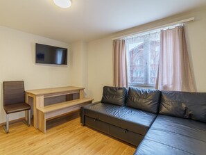 Ein kleines, möbliertes Zimmer mit einer Couch, einem Sessel, einem Holztisch und einem an der Wand befestigten Flachbildfernseher.