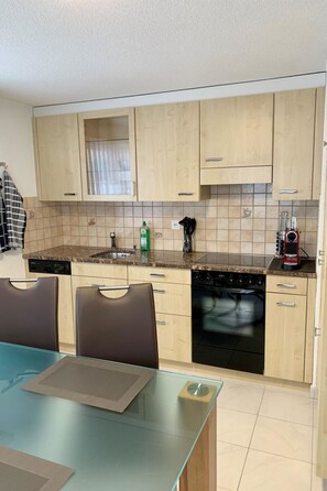 Une cuisine moderne avec des meubles en bois clair, un plan de travail en granit, un lave-vaisselle et un coin repas comprenant une table et des chaises en verre.