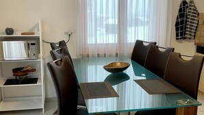 Een eetgedeelte met een tafel met glazen blad, bruine stoelen, een witte plank met keukenbenodigdheden, en een raam met transparante gordijnen.
