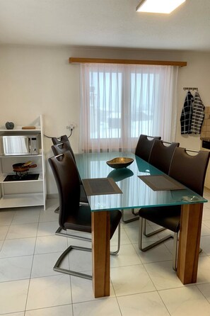 Un coin repas avec une table à plateau en verre, des chaises marron, une étagère blanche contenant des ustensiles de cuisine et une fenêtre ornée de rideaux légers.