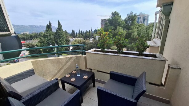 Ein Balkon mit Gartenmöbeln, einem Tisch mit Gläsern und einer Flasche sowie Blick auf Bäume und Gebäude.