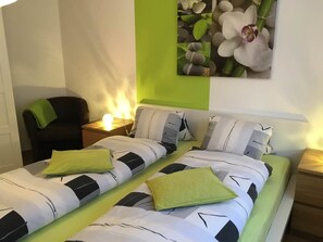 A double bed with striped bedding and two pillows, a bedside table with a lamp, a chair, and a wall-mounted picture of flowers. - Alcazar Apartments Appartement/Fewo Wohnung 3b by Interhome (Saas-Grund)