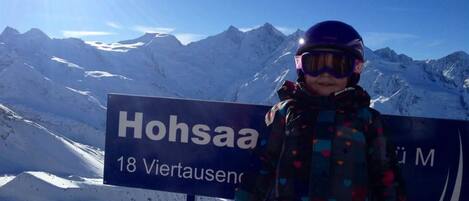A child standing in front of a sign with 'Hohsa' and '18 Viertausen' written on it, with snow-covered mountains in the background.