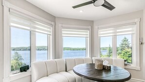 A room with a white leather sofa, a round table, and large windows with a view of a lake and trees. - Luxe Lake Front | Sauna | Pool Table | FirePit (Greater Sudbury)