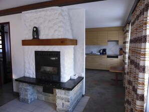 Ein Wohnzimmer mit Kamin aus Stein, einer Küche im Hintergrund und einem gemusterten Vorhang.