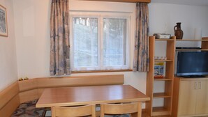 Una habitación acogedora con un banco de madera, una mesa y sillas. Hay una ventana con cortinas, un televisor sobre un mueble bajo y una estantería.