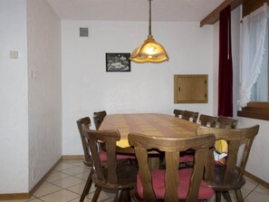 A dining area with a wooden table and chairs, a hanging light fixture, a framed picture on the wall, and a window with curtains. - Etzel Appartement/Fewo 12-Bettwohnung by Interhome (Saas-Fee)