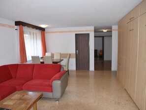 A modern living room with a red sofa, a wooden coffee table, and a dining area with a table and chairs. There is a wardrobe with multiple doors and a door leading to another room. - Acimo Appartement/Fewo 4-Bettwohnung Adam & Eva by Interhome (Saas-Fee)