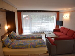 Una habitación de hotel con cama, sofá rojo, televisión y balcón con mesa y sillas.
