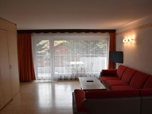 Ein Wohnzimmer mit einem roten Sofa, einem Couchtisch, einem an der Wand montierten Fernseher und Blick durch ein großes Fenster mit Vorhängen.