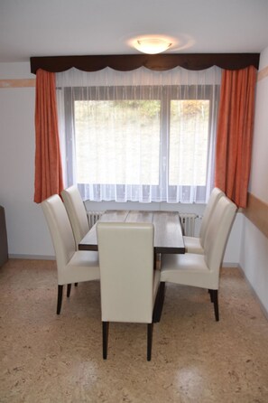 Ein Essbereich mit Tisch und vier Stühlen, ein Fenster mit Vorhängen und ein Heizkörper darunter.