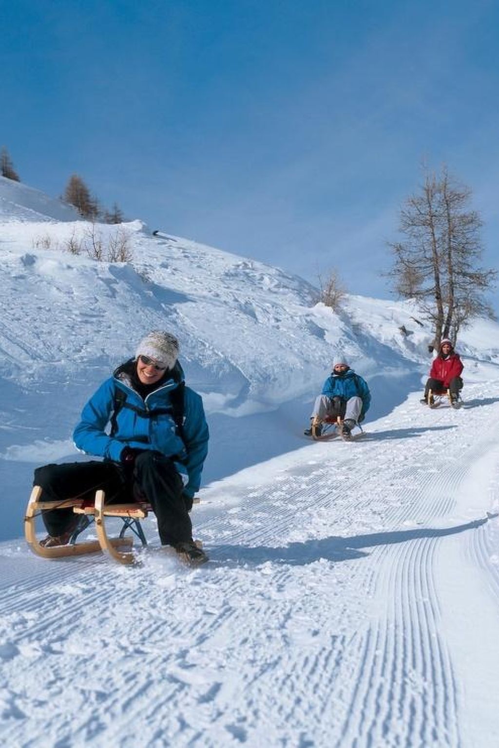 A snowy slope with three individuals on sleds, one in a blue jacket and another in a red jacket.